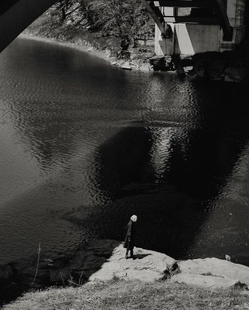 Monochromatic photograph of a person standing on a rock by a river under a bridge, creating a reflective shadow in the water.