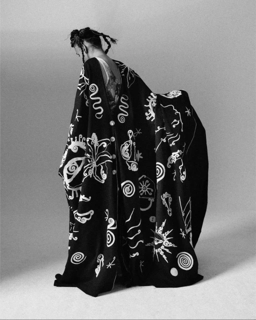 Black and white fashion shot of a person in a flowing robe with abstract, artistic patterns in a minimalist studio setting.