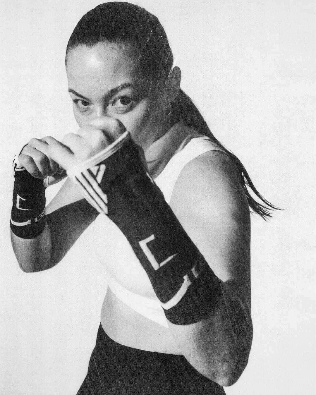 A woman with dark hair, wearing black arm wraps and athletic clothing, is in a boxing stance against a white background.