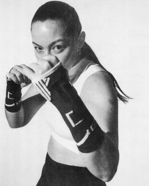 A woman with dark hair, wearing black arm wraps and athletic clothing, is in a boxing stance against a white background.