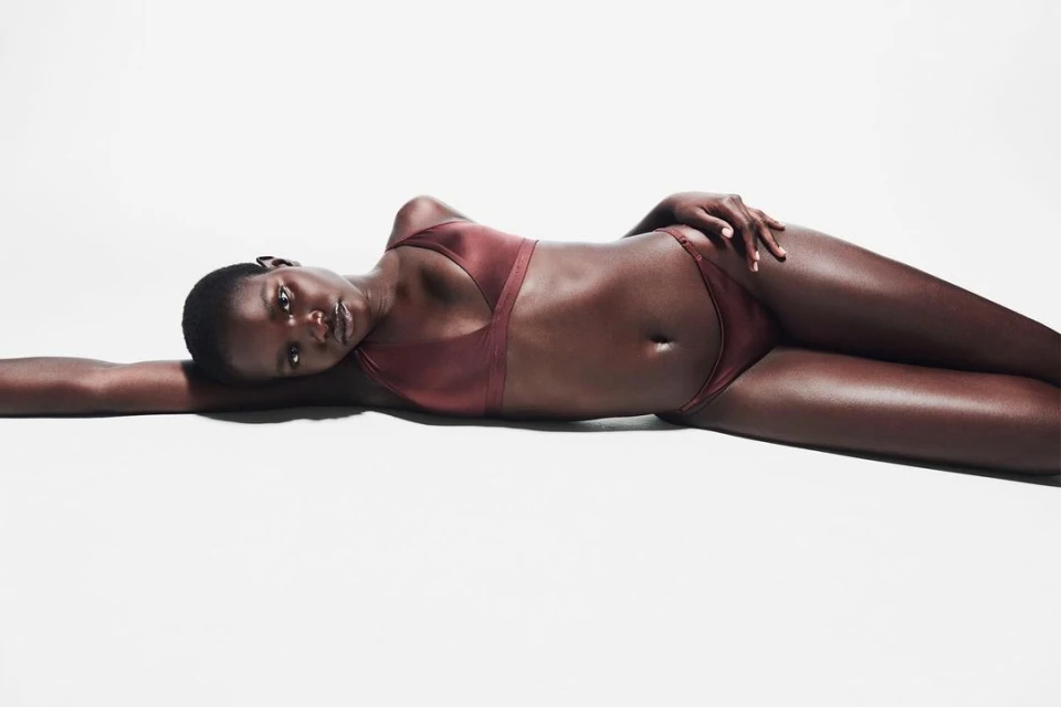 Woman with dark skin and short hair poses lying down in brown lingerie set against a white backdrop, with a serene expression.