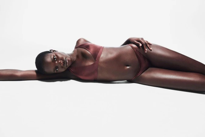 Woman with dark skin and short hair poses lying down in brown lingerie set against a white backdrop, with a serene expression.