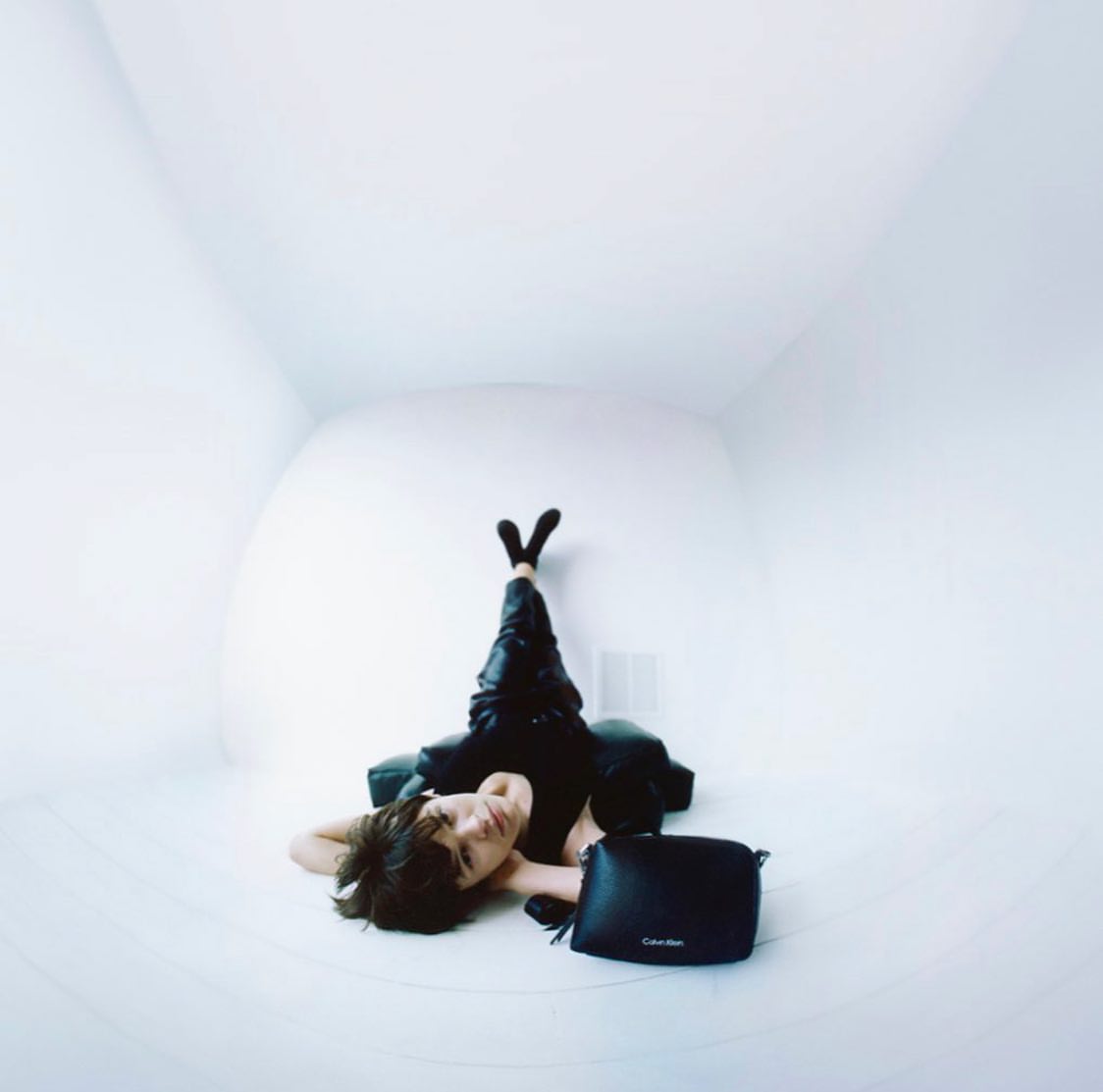 Woman in black outfit lounges in a white room with Calvin Klein handbag, creating a minimalist and high-fashion editorial aesthetic.