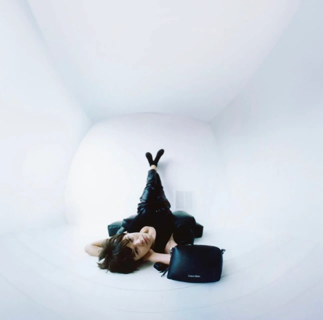 Woman in black outfit lounges in a white room with Calvin Klein handbag, creating a minimalist and high-fashion editorial aesthetic.