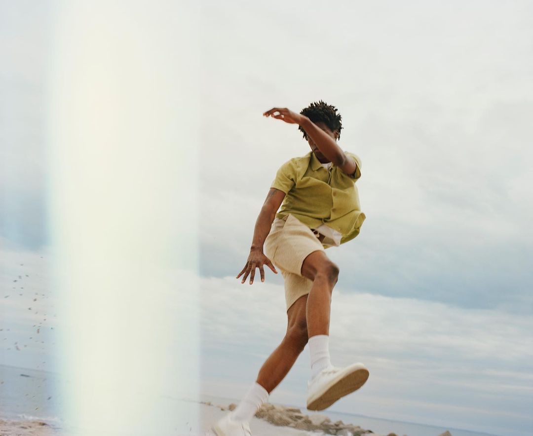 An African-American man is in mid-air on a sandy beach near the water, wearing casual clothes and looking towards the ground.