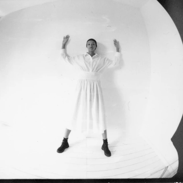Monochrome portrait of a slender adult female posing with hands up against a white wall, wearing a long white dress and black boots indoors.