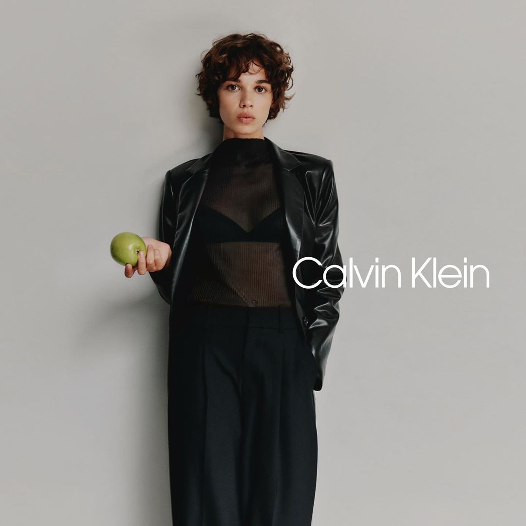 A brunette adult woman poses in a studio wearing a black jacket and holding a green apple in front of a grey background.