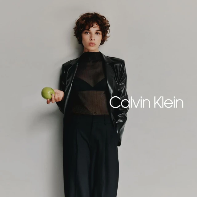 A brunette adult woman poses in a studio wearing a black jacket and holding a green apple in front of a grey background.