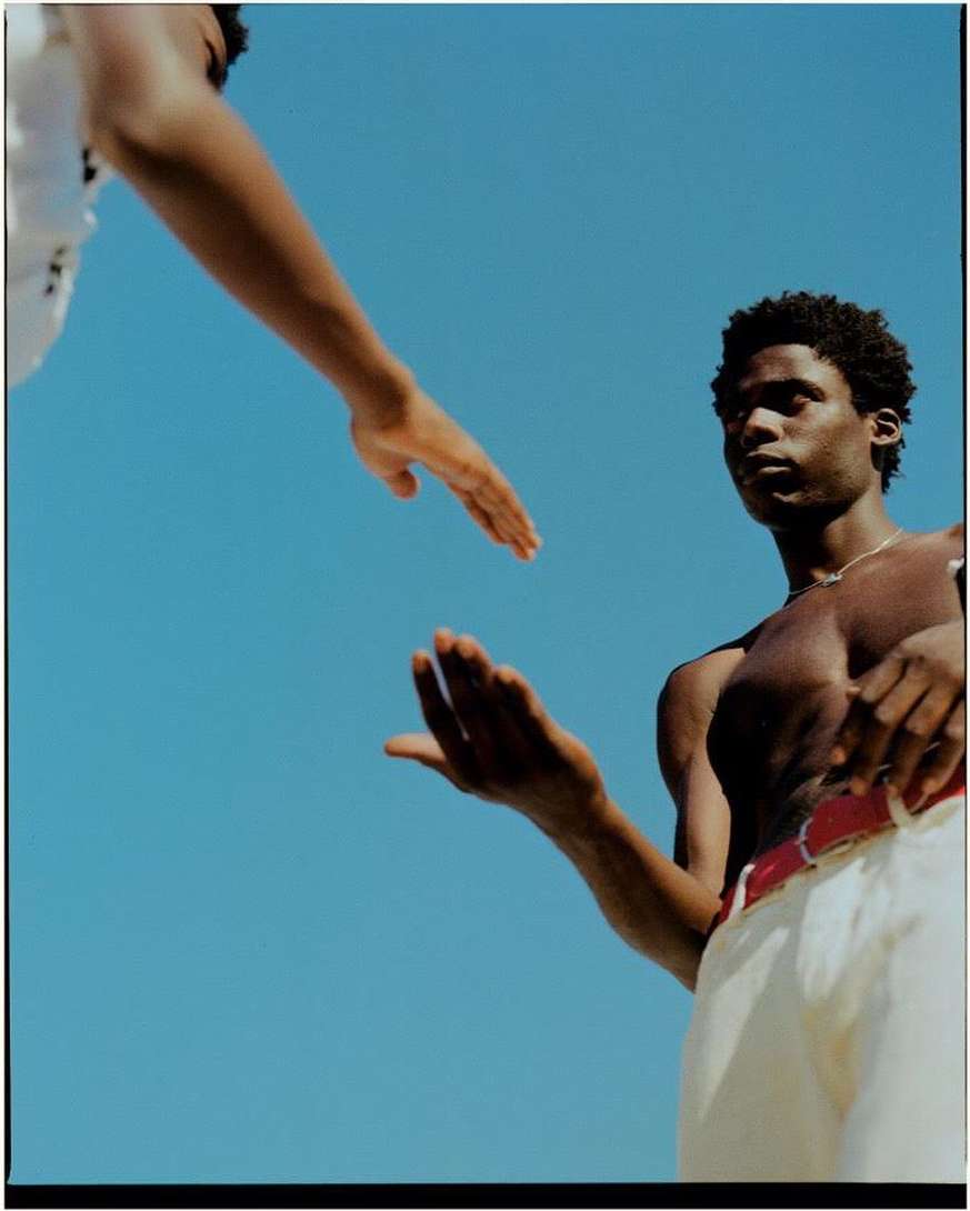 Two shirtless African American men reaching hands against a bright blue sky, wearing white pants and red belts, outdoor setting.
