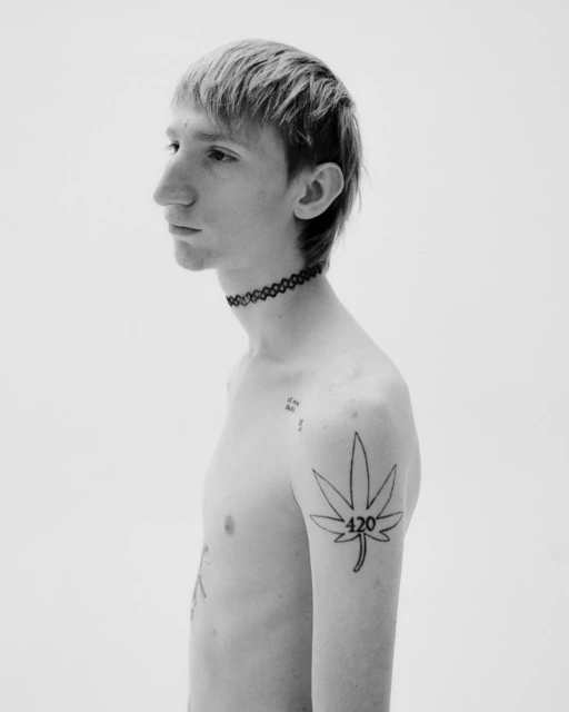 Monochrome portrait of a young adult with a tattoo on their arm, featuring a minimalist style and a neutral expression against a white backdrop.