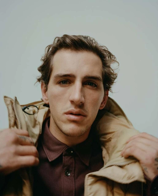 Portrait of a young adult male with brown hair wearing a tan jacket and a purple collared shirt against a plain white background.