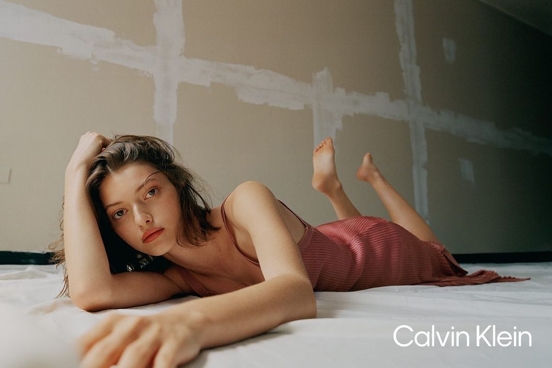 An adult woman poses in a Calvin Klein campaign, lying on a white sheet in a room with light-colored walls.