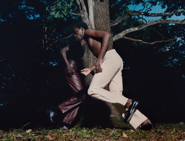 Two dark-skinned adult males walking or running outdoors against a tree, one wearing light pants, the other wearing dark pants.