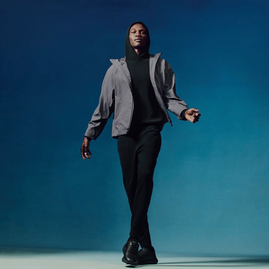 Full-length portrait of an African American male model in black athletic wear and a gray jacket, walking against a blue gradient background.
