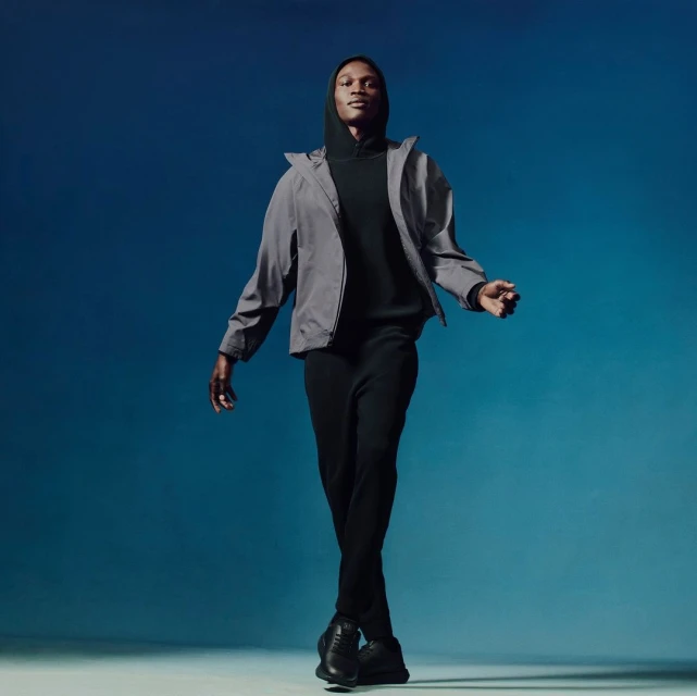 Full-length portrait of an African American male model in black athletic wear and a gray jacket, walking against a blue gradient background.