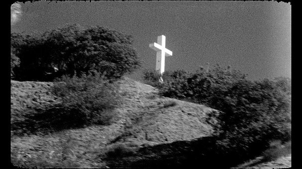 Monochrome landscape featuring a white cross atop a hill, framed by foliage, evoking a sense of stark simplicity and quiet contemplation.