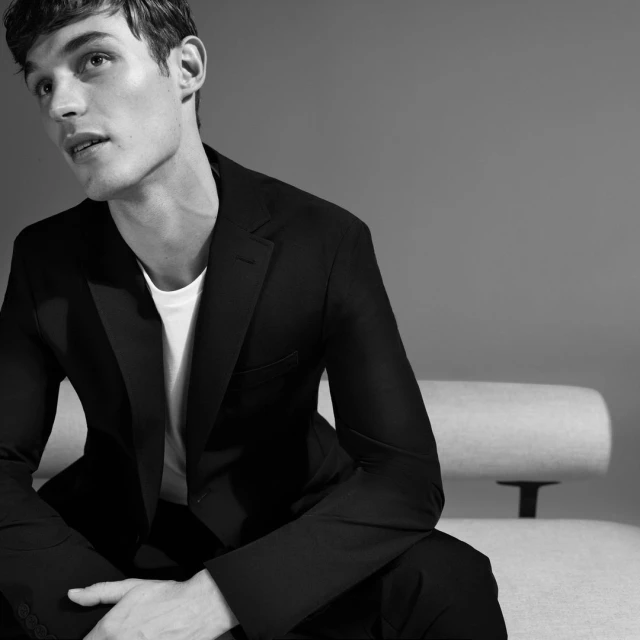 Monochrome portrait of a dark-haired adult male in a black suit and white t-shirt, seated indoors against a grey background.