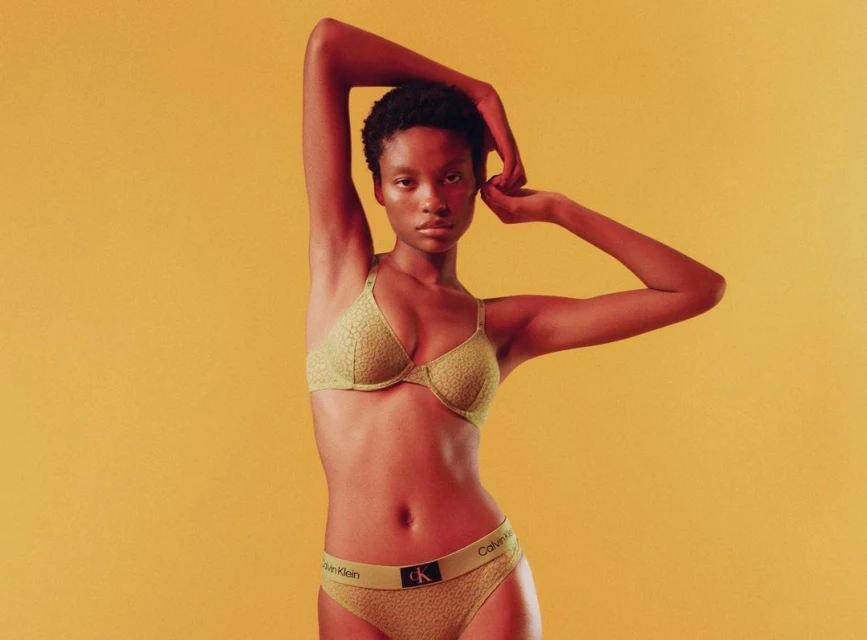 Close-up studio shot of a Black woman in a yellow bra and underwear set, posing against a solid yellow background in studio lighting.