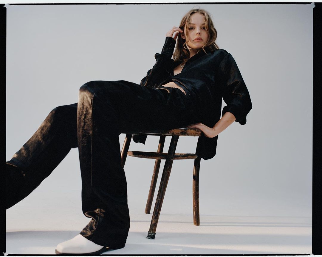 A woman in a dark silk shirt and pants sits on a wooden chair in a photo studio, posing with one hand in her hair.