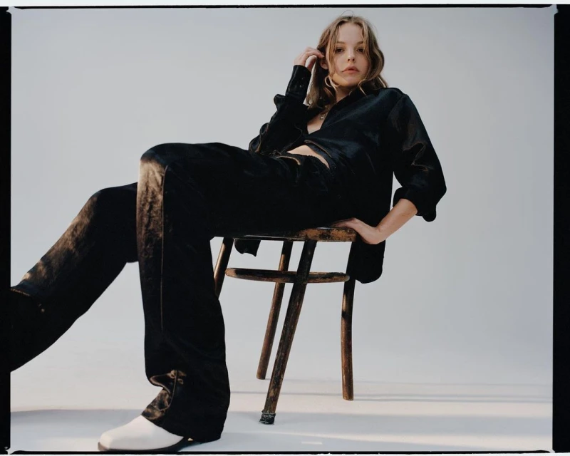 A woman in a dark silk shirt and pants sits on a wooden chair in a photo studio, posing with one hand in her hair.