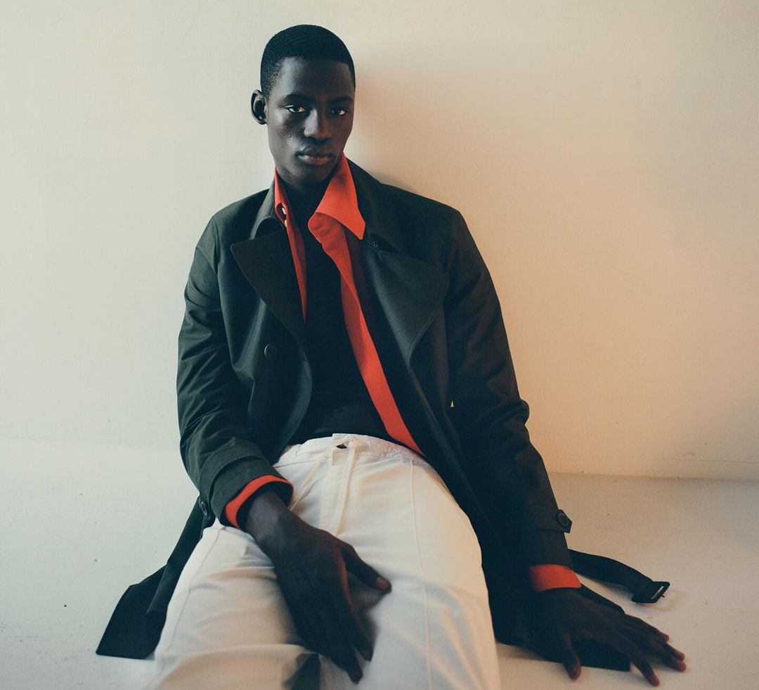 Portrait of an African male model wearing a dark trench coat and white pants, sitting on the floor against a light wall.