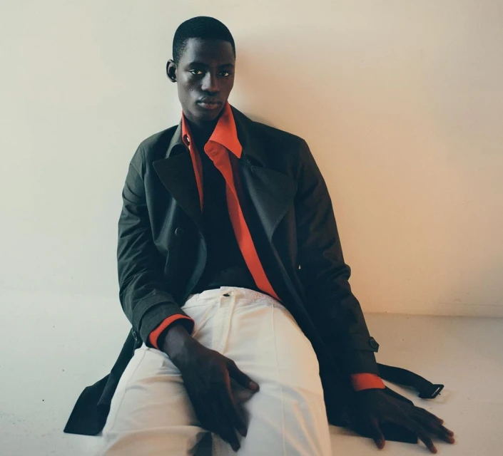Portrait of an African male model wearing a dark trench coat and white pants, sitting on the floor against a light wall.