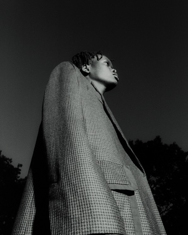 Monochrome portrait of an African-American person in a patterned coat looking up against a dark sky and silhouetted trees.