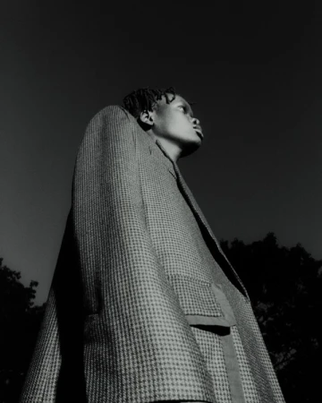 Monochrome portrait of an African-American person in a patterned coat looking up against a dark sky and silhouetted trees.