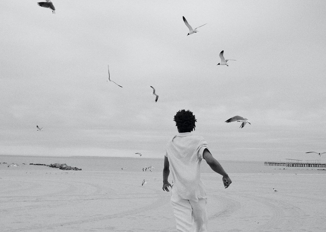 Man in white outfit faces away from the camera on a beach, surrounded by seagulls in flight against a cloudy sky in a monochrome image.