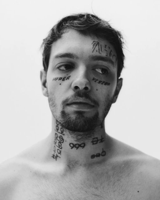 Black and white portrait of a young adult man with facial and neck tattoos, looking off-camera with a neutral expression.
