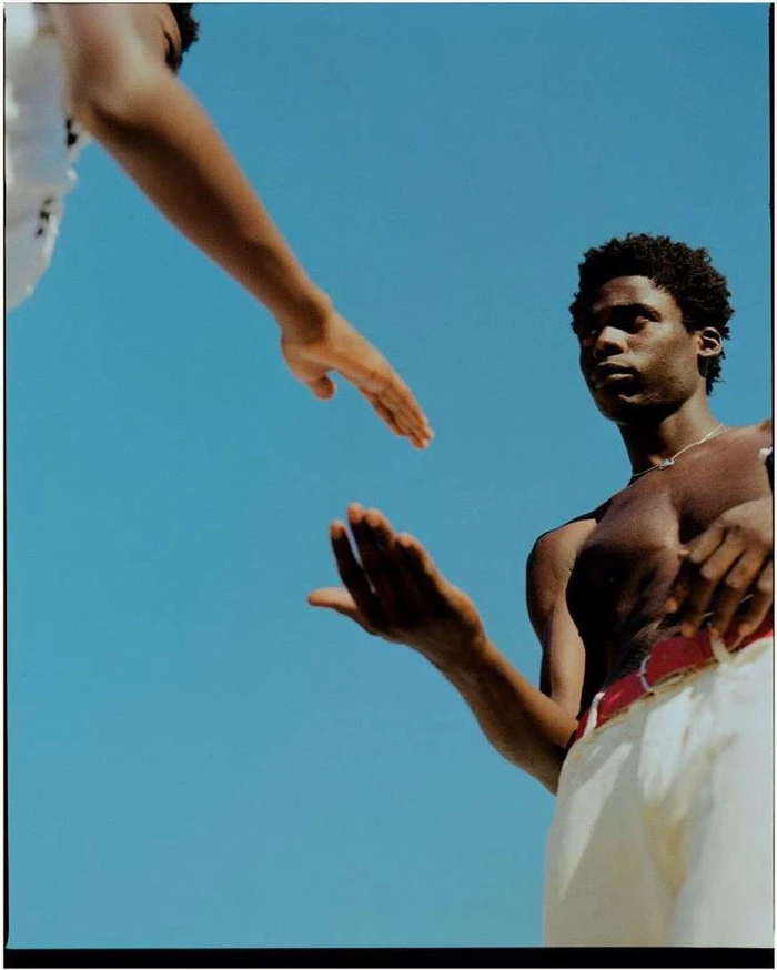 Low angle portrait of shirtless black man outdoors against a clear blue sky reaching out to touch another person's hand