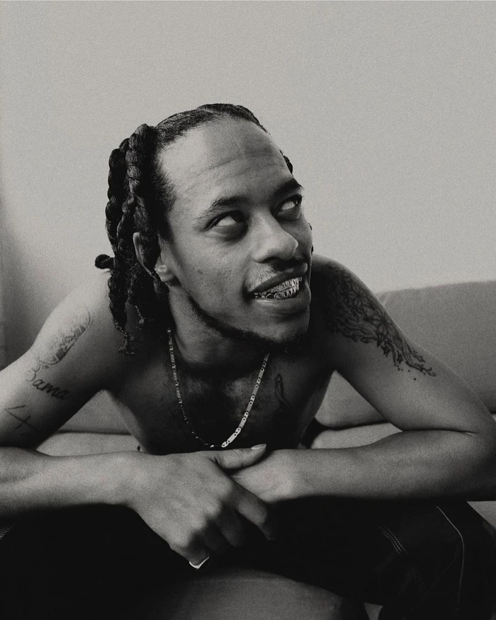 Monochrome portrait of shirtless Black man with dreadlocks and tattoos looking upward, photographed by James Brodribb.