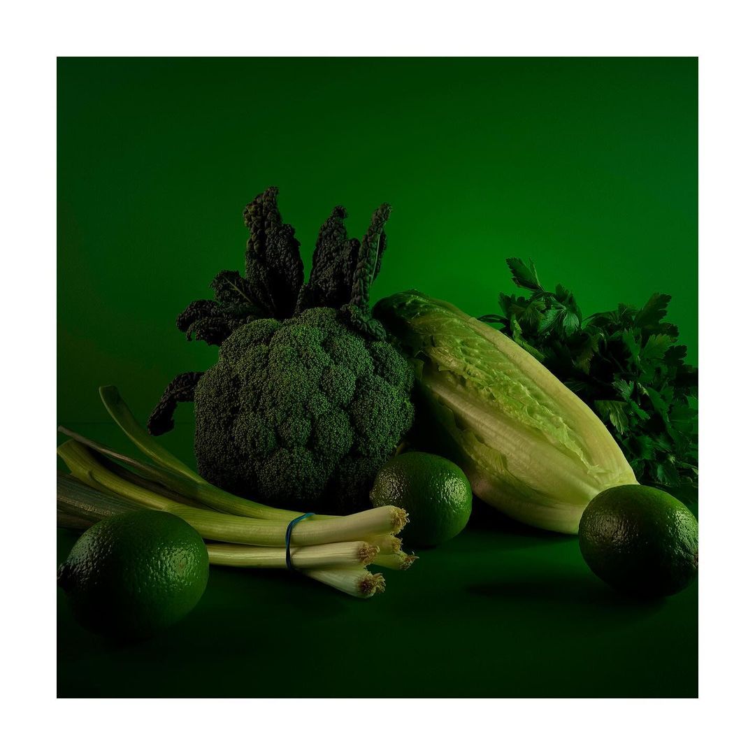 Still life featuring fresh produce: a head of broccoli, romaine lettuce, limes, green onions, parsley, and black kale, all in shades of green.