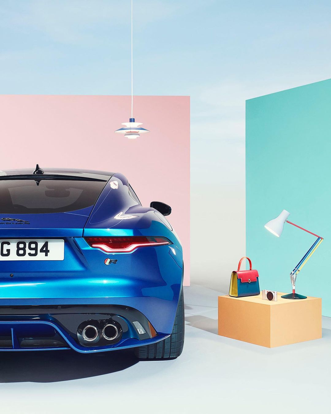 Rear view of a blue Jaguar F-Type sports car in a staged indoor setting with geometric shapes and a designer handbag and lamp.