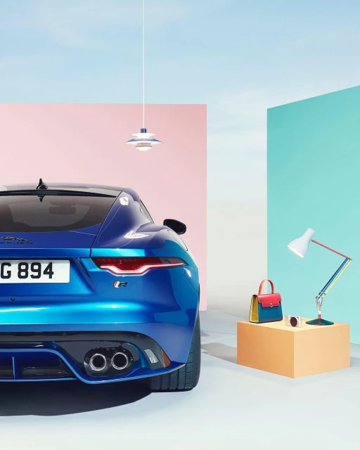 Rear view of a blue Jaguar F-Type sports car in a staged indoor setting with geometric shapes and a designer handbag and lamp.