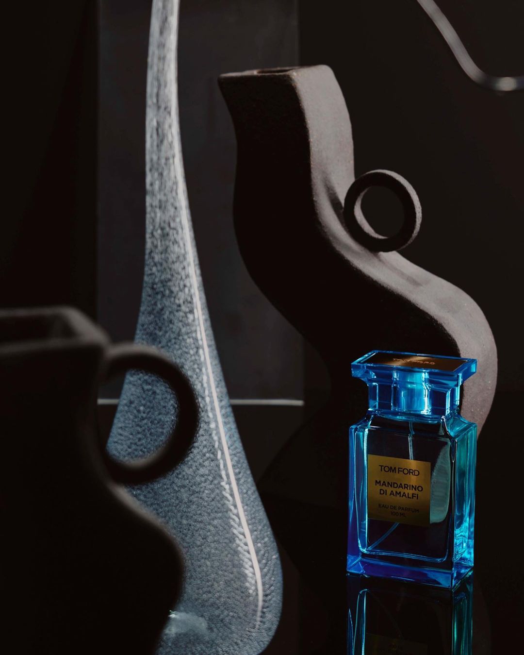 Still life shot of a blue perfume bottle with glass vases and a black ceramic jug, captured in a dark, moody studio setting.