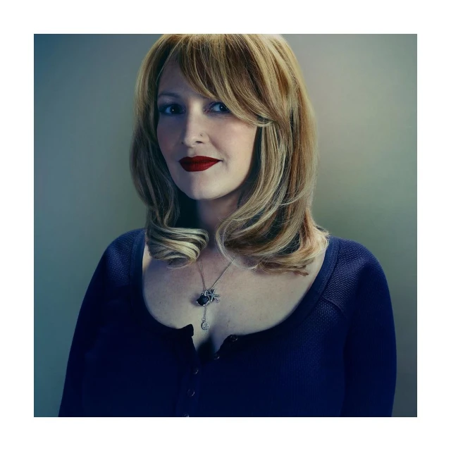 Portrait of a blonde woman with red lipstick wearing a navy blue top and silver necklace against a light-beige studio backdrop.