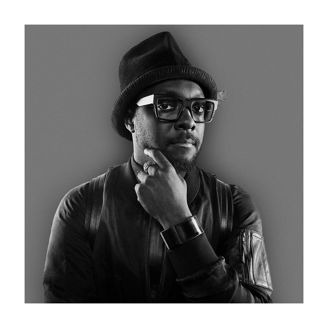 Portrait of Will.i.am wearing a black hat and eyeglasses, posing in a studio with a monochrome color palette.