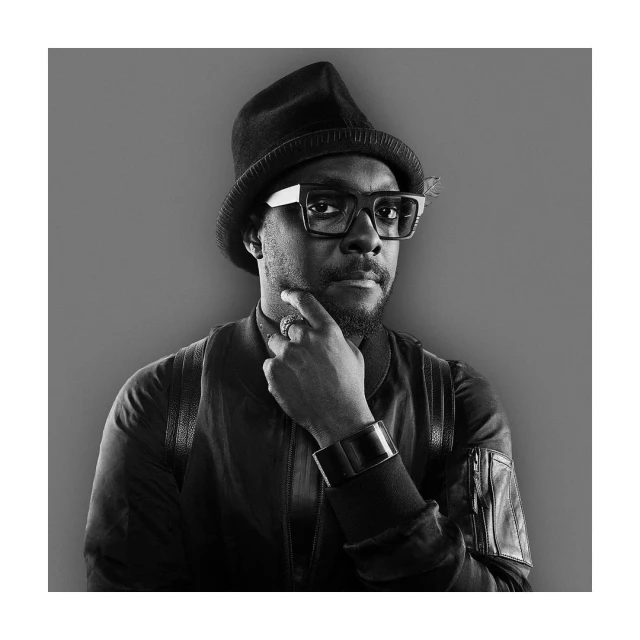 Portrait of Will.i.am wearing a black hat and eyeglasses, posing in a studio with a monochrome color palette.