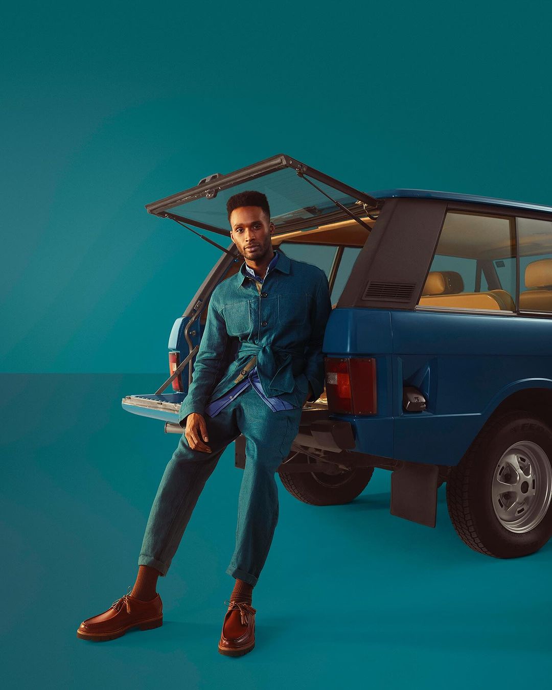 Adult male fashion model wearing blue denim suit posing with classic blue SUV with open hatchback door in a studio setting.