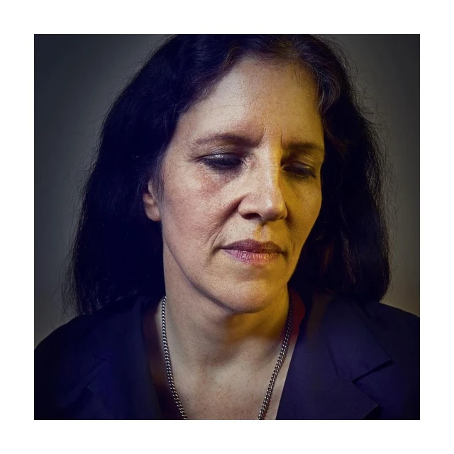 Portrait of a middle-aged woman with dark hair looking down, wearing a dark jacket and silver necklace against a grey background.