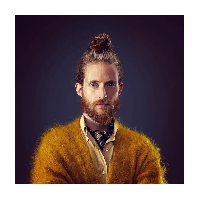 Portrait of a bearded adult male with hair in a bun, wearing a yellow shirt and cardigan, and a patterned cravat, against a dark background.