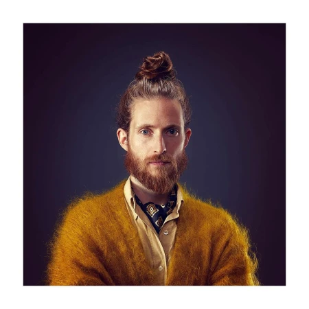 Portrait of a bearded adult male with hair in a bun, wearing a yellow shirt and cardigan, and a patterned cravat, against a dark background.