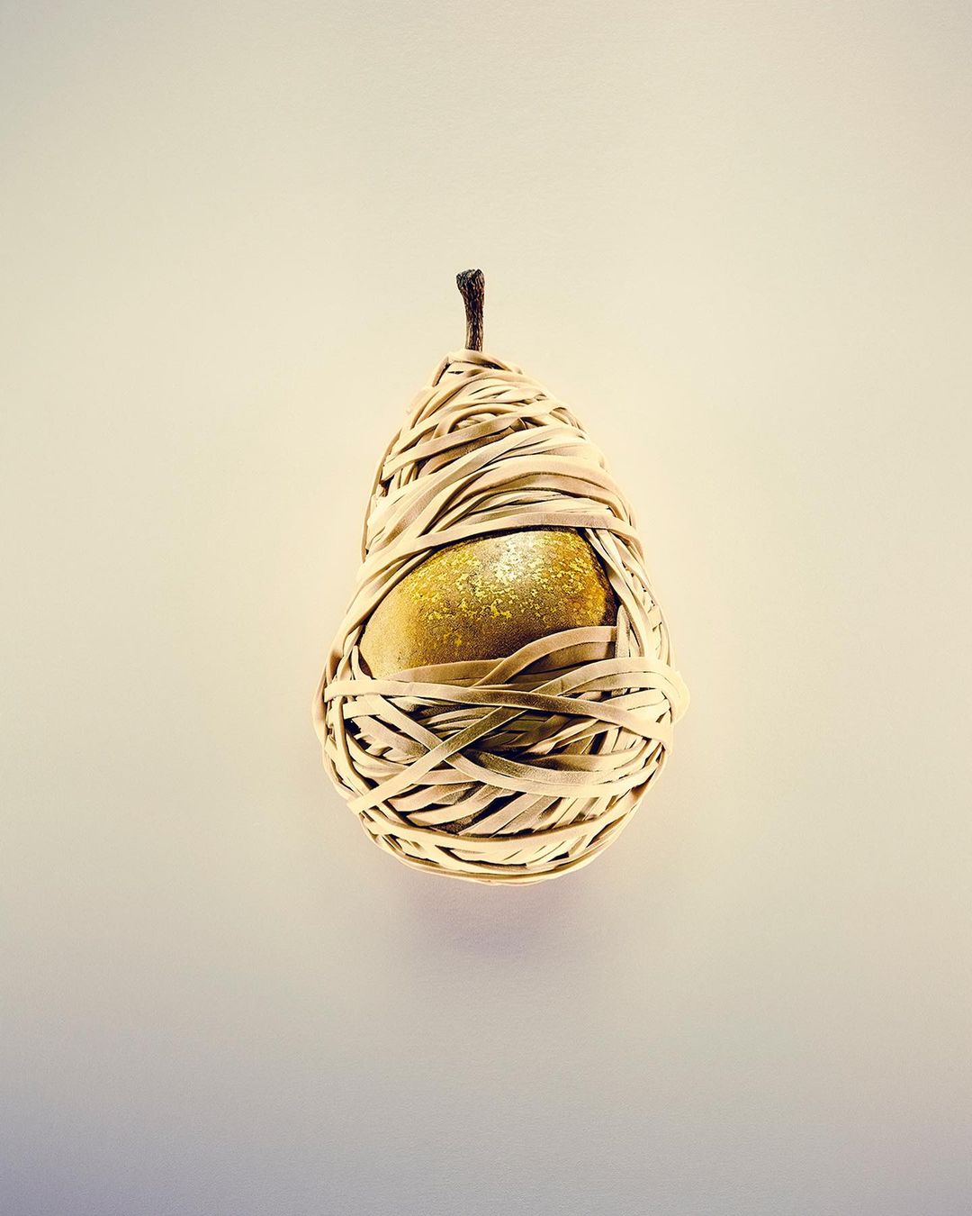 Golden pear wrapped with tan paper strips on a light yellow background in a studio setting. Food and product photography.