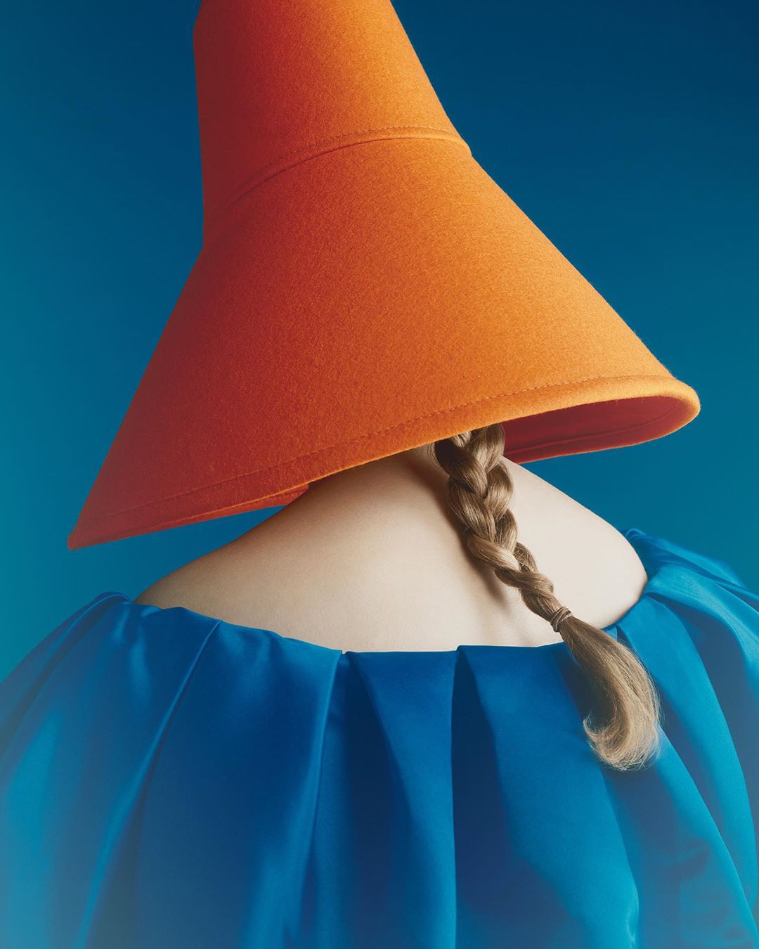 Woman wearing a conical orange hat and blue dress stands against a vibrant blue background in this minimalist studio fashion shot.