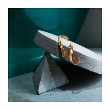 Elegant gold ribbon-shaped earrings are displayed atop geometric concrete blocks against a muted blue and teal abstract backdrop.