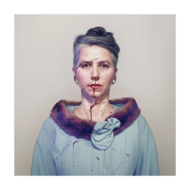 Woman with gray hair and a light blue jacket with fur collar looking at the camera. Dark liquid or paint is smeared on her face.