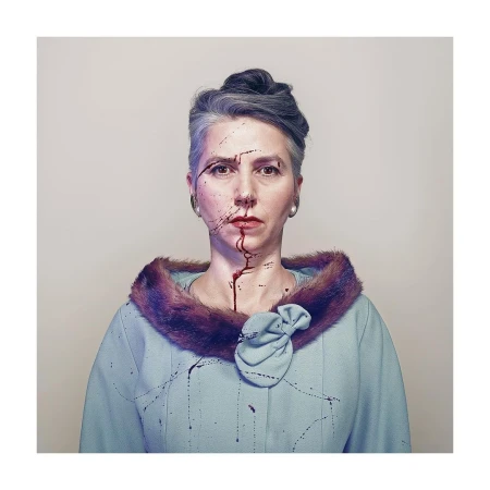 Woman with gray hair and a light blue jacket with fur collar looking at the camera. Dark liquid or paint is smeared on her face.
