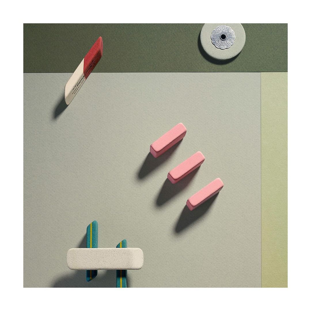 Still life of erasers in various shapes and colors arranged on a flat surface with muted green, yellow, and gray tones, lit from above.