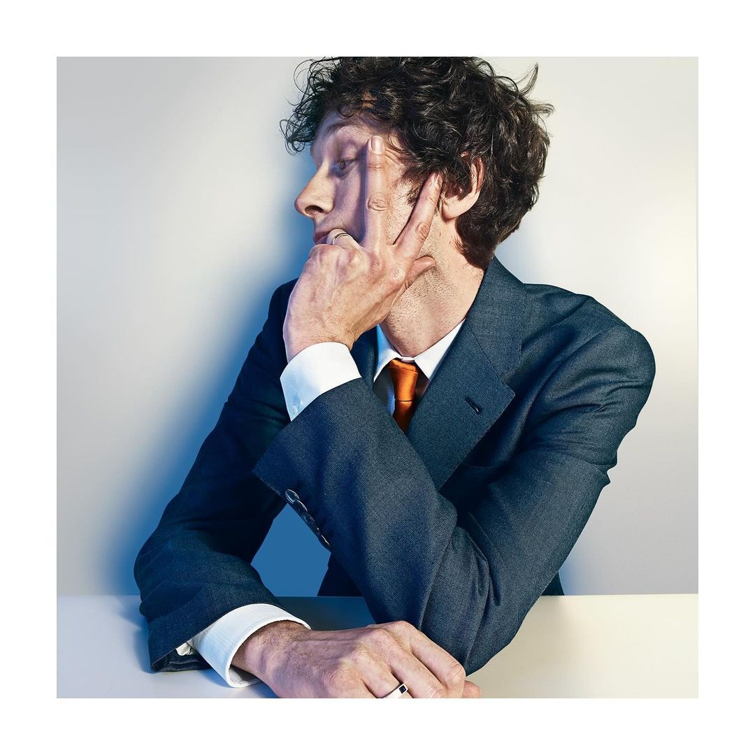 Adult male in business suit poses in a photo studio with his fingers on his face, wearing an orange tie. Head and shoulder shot.